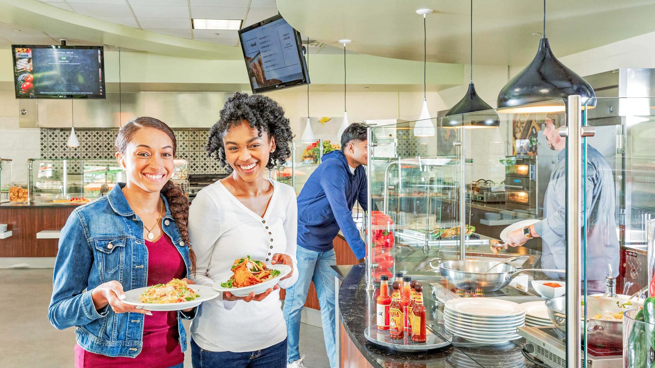 Two students holding dishes and smiling while standing in cafeteria